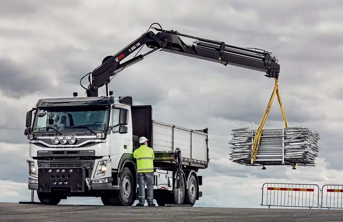 Camion équipé d'une grue auxiliaire de chargement levant un lot de barrières, opérateur au sol en tenue haute visibilité